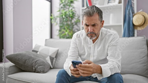 Upset young hispanic man, sporting grey hair and a beard, engrossed in a serious smartphone ordeal while comfortably nestled on his living room sofa at home