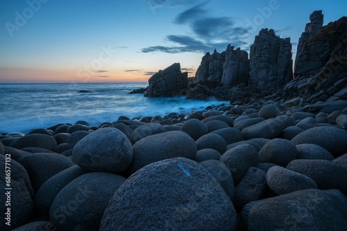 Beach shore with large rocks and crashing waves illuminated by the warm glow of the sunset sky © Wirestock