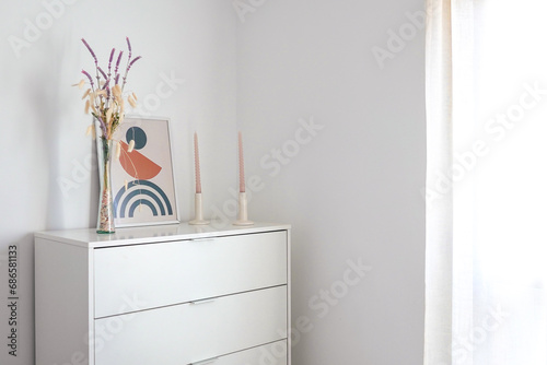 comoda de habitación blanca , con flores, velas y un cuadro