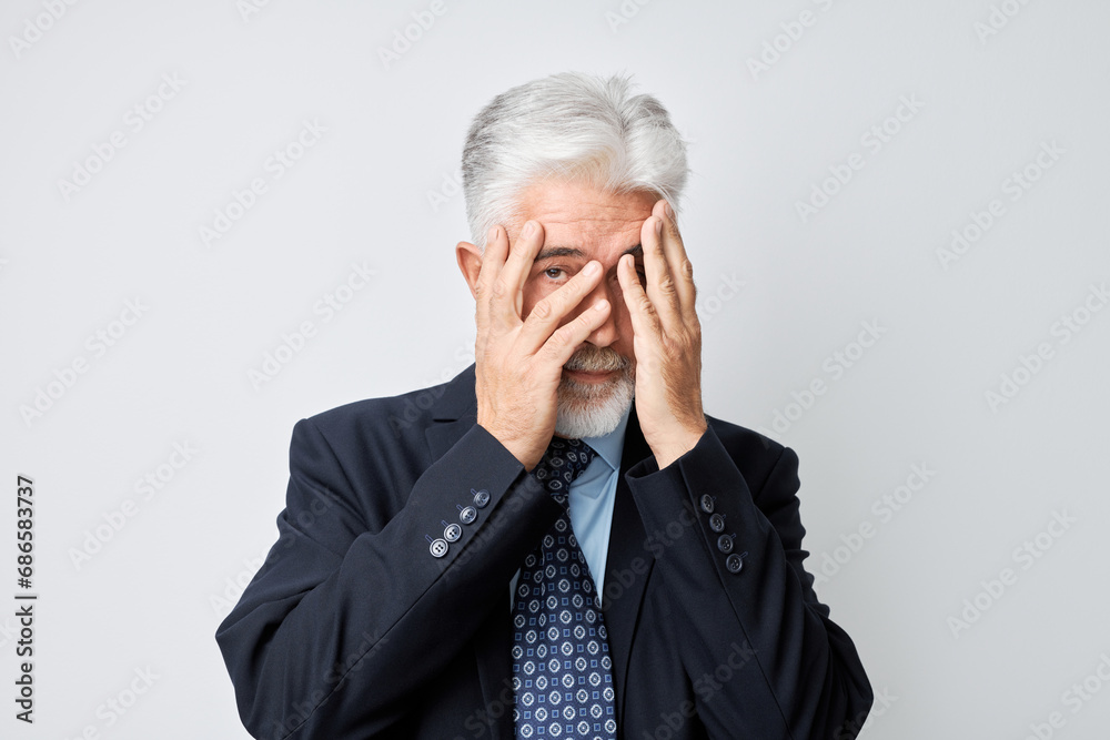 Portrait of elderly man covering eyes with hand, peeking through fingers isolated on white studio background.