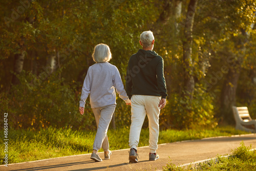Married senior couple walking together on a sunny summer evening. From behind an old man and woman in sports clothes holding hands walking on a park walkway among green trees. Exercise, motion concept photo