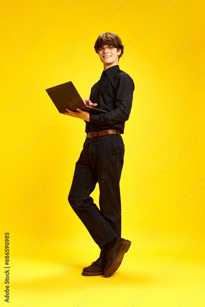 Study and work online from anywhere in world. Full length portrait of young attractive man posing with laptop against vivid yellow studio background.