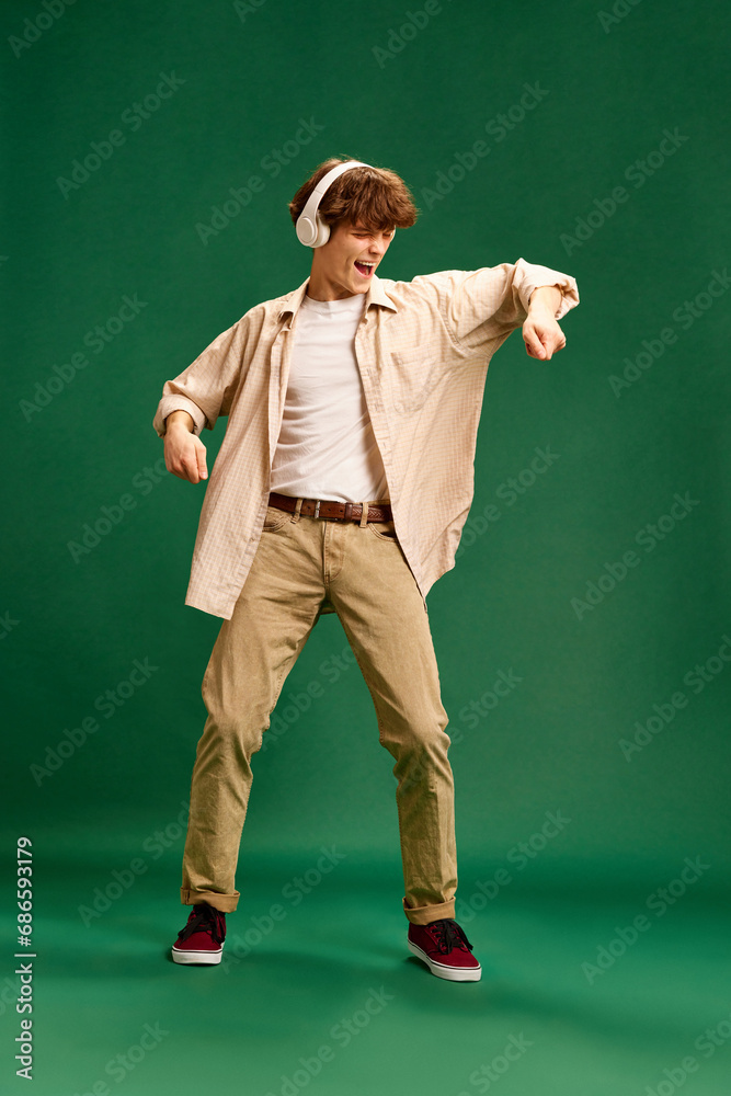 Full-length portrait of emotional young man wearing smart casual retro outfit dancing in headphones against green studio background.