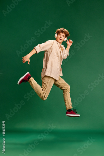 Full length portrait of young man, student dressed retro fashion outfit in headphones jumps high against green studio background.