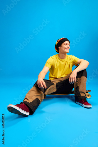 Positive, smiling man in sunglasses wearing in rastaman, reggie style and posing sitting on skateboard against blue background.