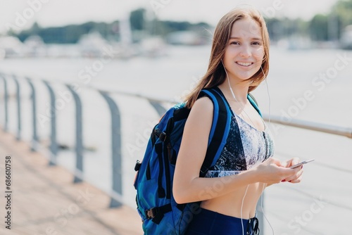 Smiling athlete with backpack and headphones using phone on sunny pier