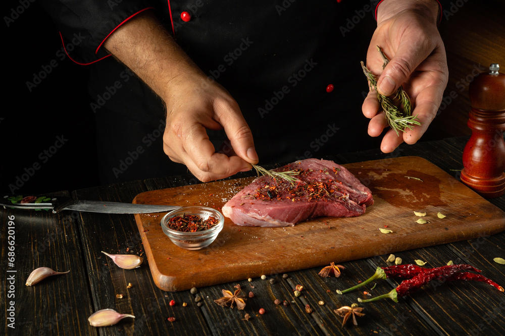 The chef adds dry aromatic rosemary to the meat steak. The process of cooking veal meat or shish kebab on the kitchen table with spices and salt