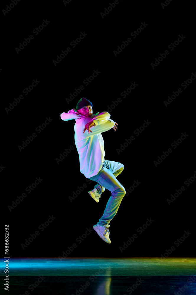 Modern dance. Breakdance male dancer, athletic guy dancing freestyle in stylish outfit against black background in neon light.