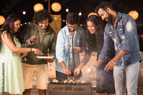 Group of friends busy cooking barbecue grills for dinner during night party - concept of togetherness, birthday gathering and celebration photo