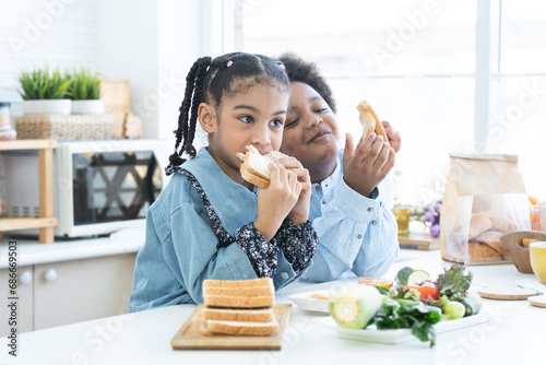 African little child girl eating homemade sandwiches in hands, standing in kitchen at home with brother, prepare food for breakfast. Siblings, kids make sandwich. Selective focus on girl