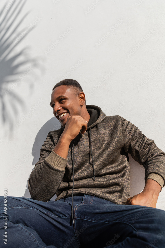satisfied african american man in jeans and hoodie sitting near white wall satisfied african american man in jeans and hoodie sitting near white wall