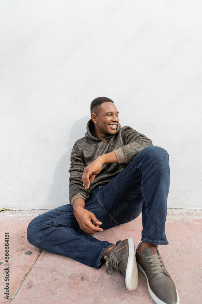 cheerful african american man in jeans and hoodie smiling while sitting near white wall