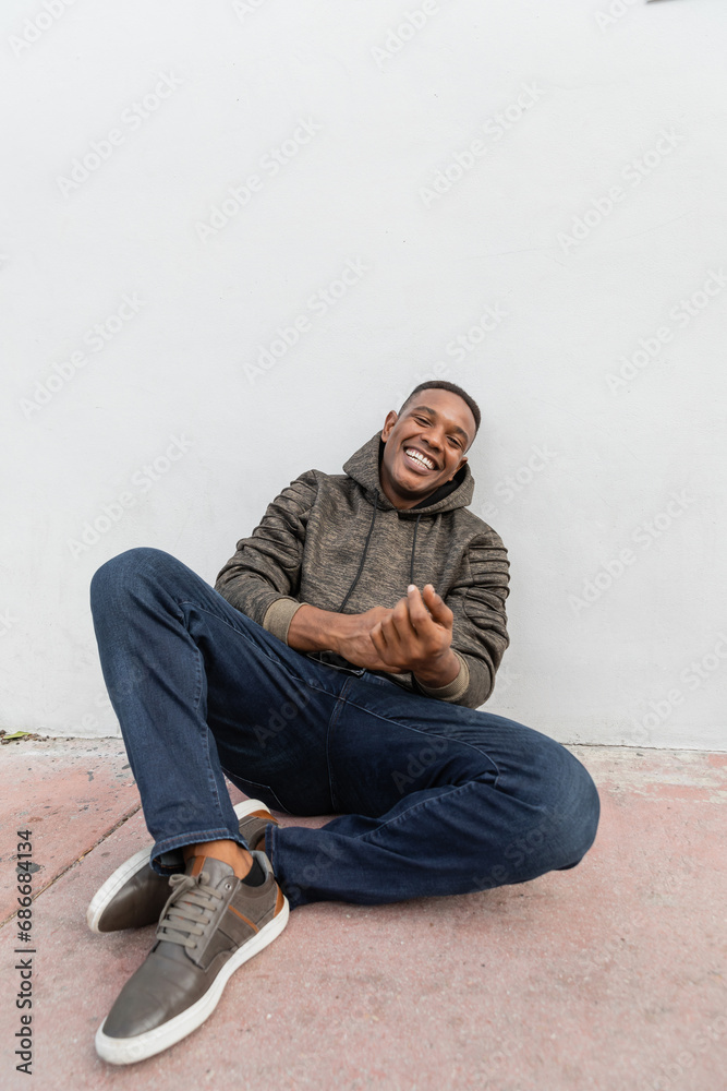 happy african american man in jeans and hoodie smiling while sitting near white wall