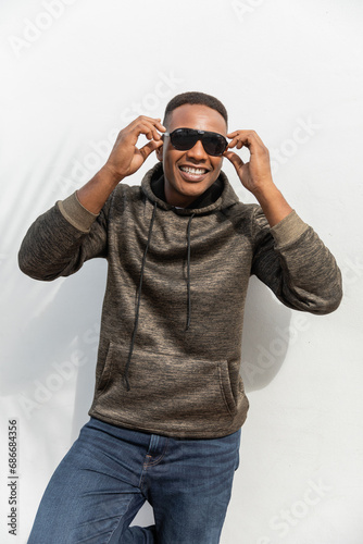 happy african american man in hooded sweatshirt wearing sunglasses near white wall © LIGHTFIELD STUDIOS