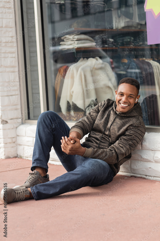 full length of cheerful african american man in hoodie sitting near showcase of shop in Miami