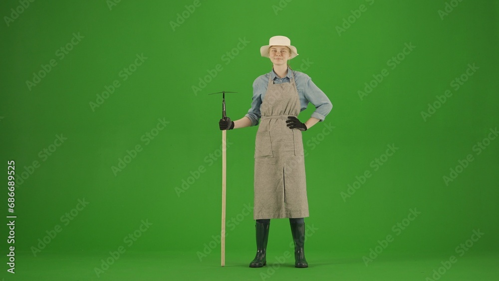 Portrait of farmer in working clothing on chroma key green screen. Gardener woman standing posing at camera with hoe, smiling face. Portrait of farmer in working clothing on chroma key green screen. Gardener woman standing posing at camera with hoe, smiling face.