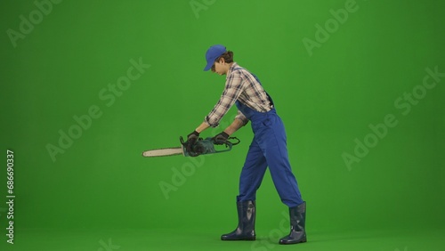 Portrait of farmer in working clothing on chroma key green screen. Gardener standing and cutting wood with chainsaw.