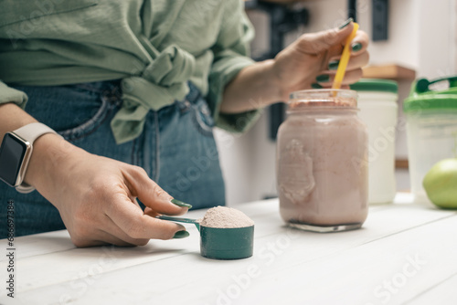Young woman in jeans and shirt holding measuring spoon with protein powder, glass jar of protein drink cocktail, milkshake or smoothie above white wooden table with chocolate pieces, bananas, apples photo