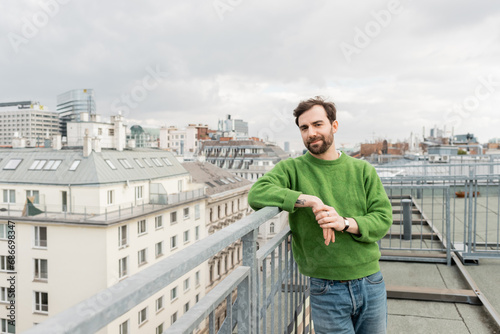 handsome man in green jumper looking away while standing on rooftop terrace in Vienna, Austria