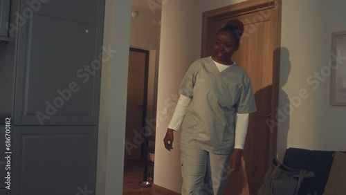 Medium shot of young African American health worker in grey uniform coming home from night shift to family, hugging son browsing on smartphone in kitchen, and kissing husband