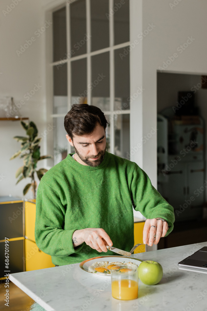 bearded man in green jumper holding cutlery near breakfast in modern kitchen at home bearded man in green jumper holding cutlery near breakfast in modern kitchen at home