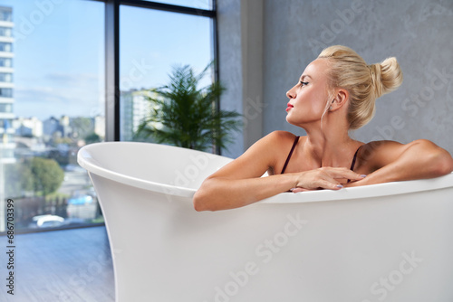Gorgeous blond caucasian lady looking at window with scenic view, while leaning on edge of bathtub. Side view of pensive woman with silky skin, taking bath at home. Relax and recreation concept. 