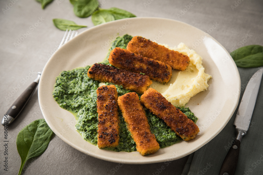 Gebratene Fischstäbchen mit Rahmspinat und Kartoffelpüree auf Teller mit Besteck, Serviette, Spinat Blätter roh und hell grau Stein Beton Hintergrund Gebratene Fischstäbchen mit Rahmspinat und Kartoffelpüree auf Teller mit Besteck, Serviette, Spinat Blätter roh und hell grau Stein Beton Hintergrund
