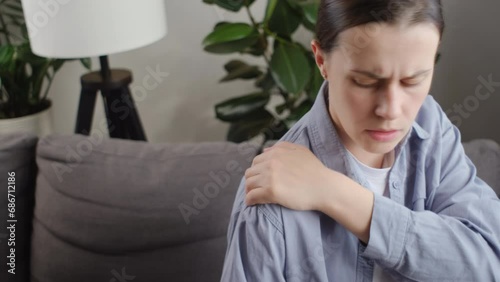 Close up of upset sad young lady sitting on sofa doing massage in shoulder with other hand. Arm injury, shoulder and forearm pain, people with body muscle problems, healthcare and medicine concept photo