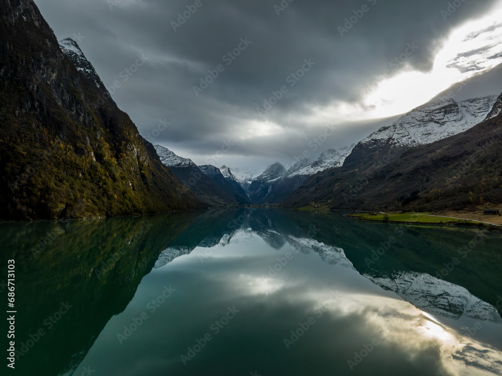 aerial view over Oldevatn lake with cyan water surrounded by mountains reflected in water with snowy peaks and with sun breaking through the clouds