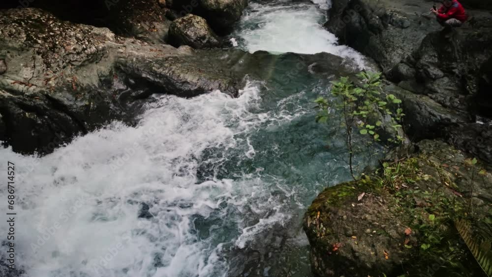 Man is sitting on the shore of forest river in mountains of Georgia and taking photo of it on his smartphone. Slow motion of male tourist trekking to Mingrelia woodland and admiring majestic cascade Man is sitting on the shore of forest river in mountains of Georgia and taking photo of it on his smartphone. Slow motion of male tourist trekking to Mingrelia woodland and admiring majestic cascade