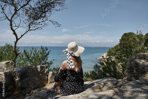 Girl in hat and polka dot dress sits on seashore and looks into distance. Salou, Costa Dorada in province of Tarragona. Spain.