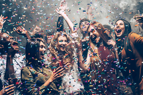 Group of joyful young friends dancing and enjoying celebration in night club while confetti flying around