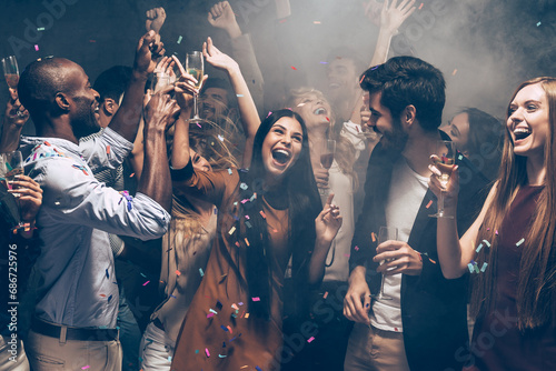 Group of cheerful young people dancing and enjoying celebration in night club while confetti flying around