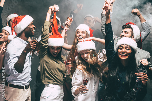 Happy young people in Christmas hats dancing and celebrating New Year in night club while confetti flying around