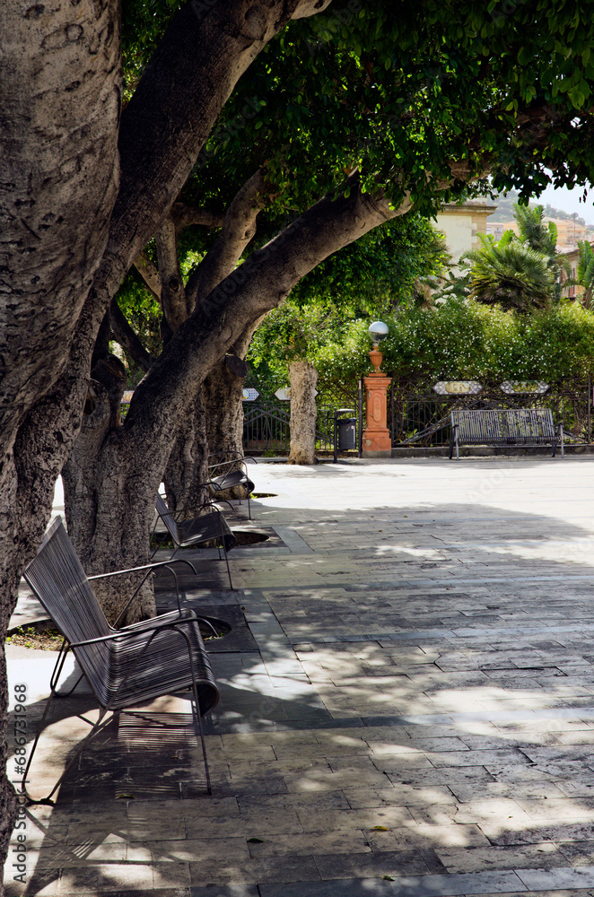 Close-up view pedestrian cobblestone alley with vintage metal benches that are in the shade of green leaves trees. Typical Sicilian cityscape of small village Santo Stefano di Camastra