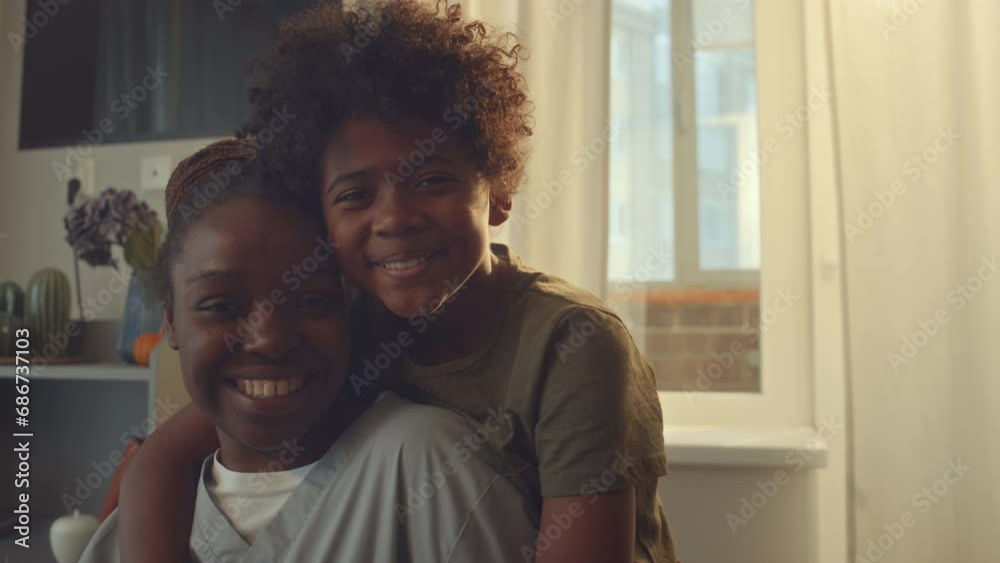 Medium close-up portrait shot of young single African American mother in health worker uniform posing in kitchen with 10-year-old son, who is hugging her, looking at camera and smiling Medium close-up portrait shot of young single African American mother in health worker uniform posing in kitchen with 10-year-old son, who is hugging her, looking at camera and smiling