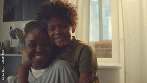 Medium close-up portrait shot of young single African American mother in health worker uniform posing in kitchen with 10-year-old son, who is hugging her, looking at camera and smiling photo