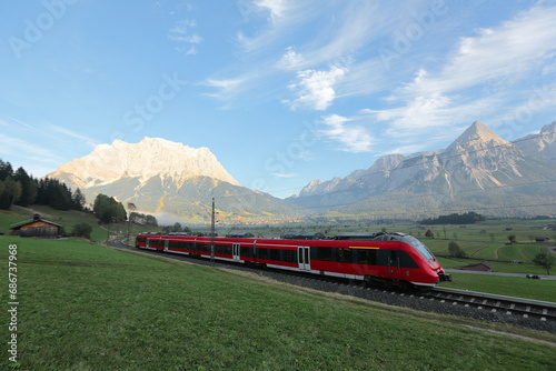 Panoramic view of a train traveling on green fields with Mountain Zugspitze in background on a beautiful sunny day in Lermoos, Tirol, Austria ~ Beautiful summer scenery of idyllic Tyrolean countryside