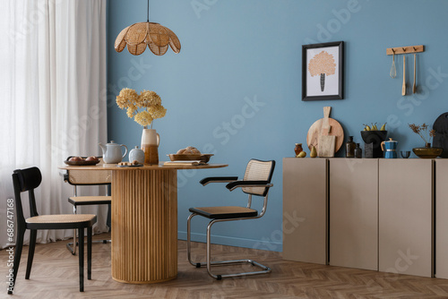 Warm and cozy dining room interior with mock up poster frame, round table, rattan chairs, beige commode, lamp, vase with flowers, kitchen cutboard and personal accessories. Home decor. Template.