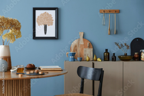 Dinning room and kitchen interior with mock up poster frame, round table, rattan chair, beige commode, vase with dried flowers, cup, pitcher and kitchen accessories. Home decor. Template.