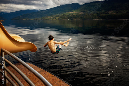 Boy jumping out of the end of a waterslide on a houseboat on a fall day on Shuswap Lake; British Columbia, Canada