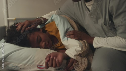 Medium close-up shot of young black boy asleep in bedroom, female doctor mom in hospital gown walking in, tucking duvet, stroking his hair and kissing goodnight, after returning late from shift