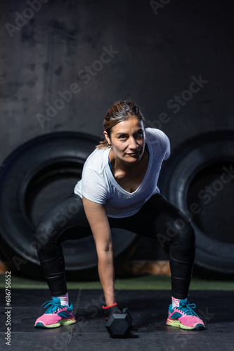 Middle-aged Latina woman with a dumbbell in a gym with a black background and two tires, new year's resolutions