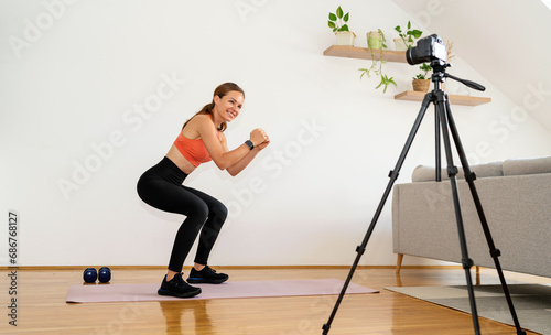 Female person doing sports training at home, fitness exercise squats at home in front of video camera on tripod.