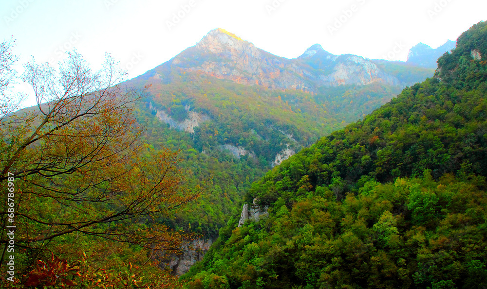 Classic view from Eremo di Soffiano near Sarnano at the massive rocky crests of the Sibillini Mountains interspersed with dense thickets, timid sunlight, vegetation-filled slope and autumn foliage