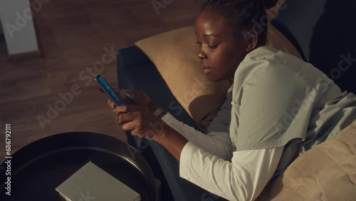Medium top shot of black female hospital worker in uniform sleeping on couch at home, alarm clock beeping on smartphone, woman waking and getting up to go to shift photo