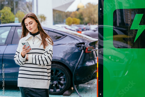Eco friendly rechargeable EV car concept. Young woman in strip sweater standing with smart phone, waiting for her electric car to charge on a public charging station outdoors.
