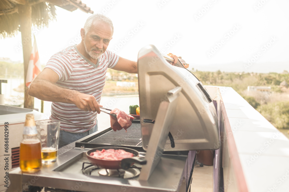 Mature man grilling steaks on a gas grill on his penthouse terrace - obrazy, fototapety, plakaty 