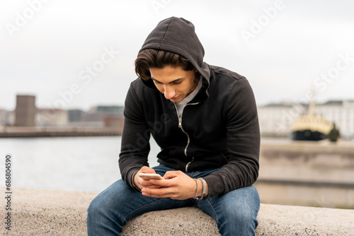 Canvas Print Denmark, Copenhagen, young man sitting on wall at the waterfront using cell phon