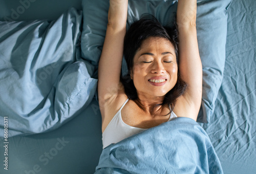 Happy asian woman wakes up well rested stretches arms in bed photo